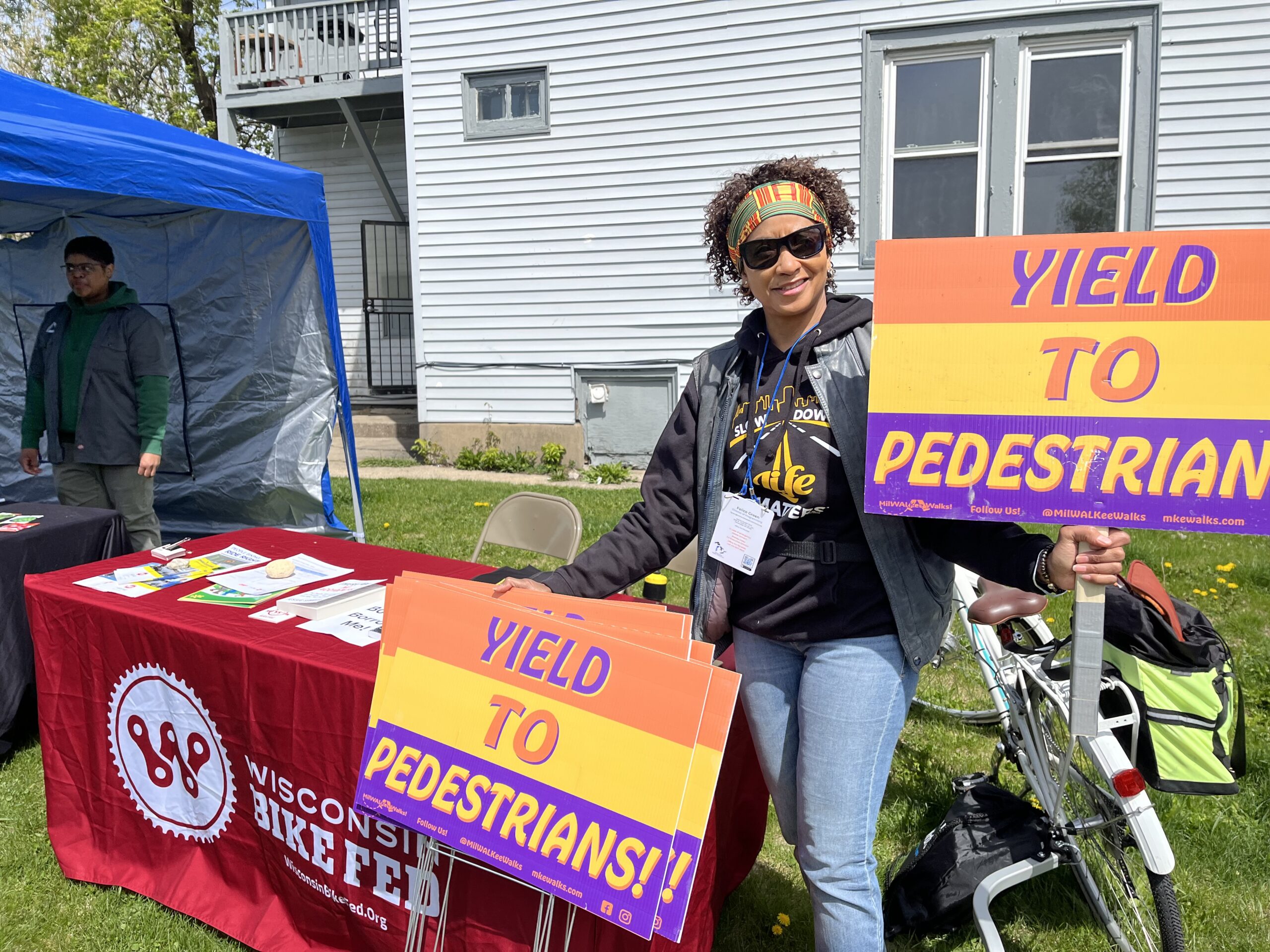 Woman holding yard signs saying "Yield to Pedestrians." There is a bike, table, and tent in the background.