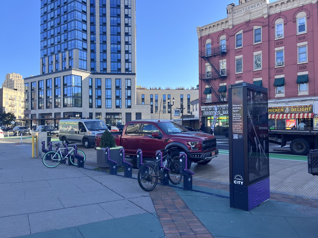 There are tall buildings in the background as well as cars parked along a street. In the foreground, there is a series of bike racks that have purple arms for users to lock their bikes to the rack without bringing a private lock. 