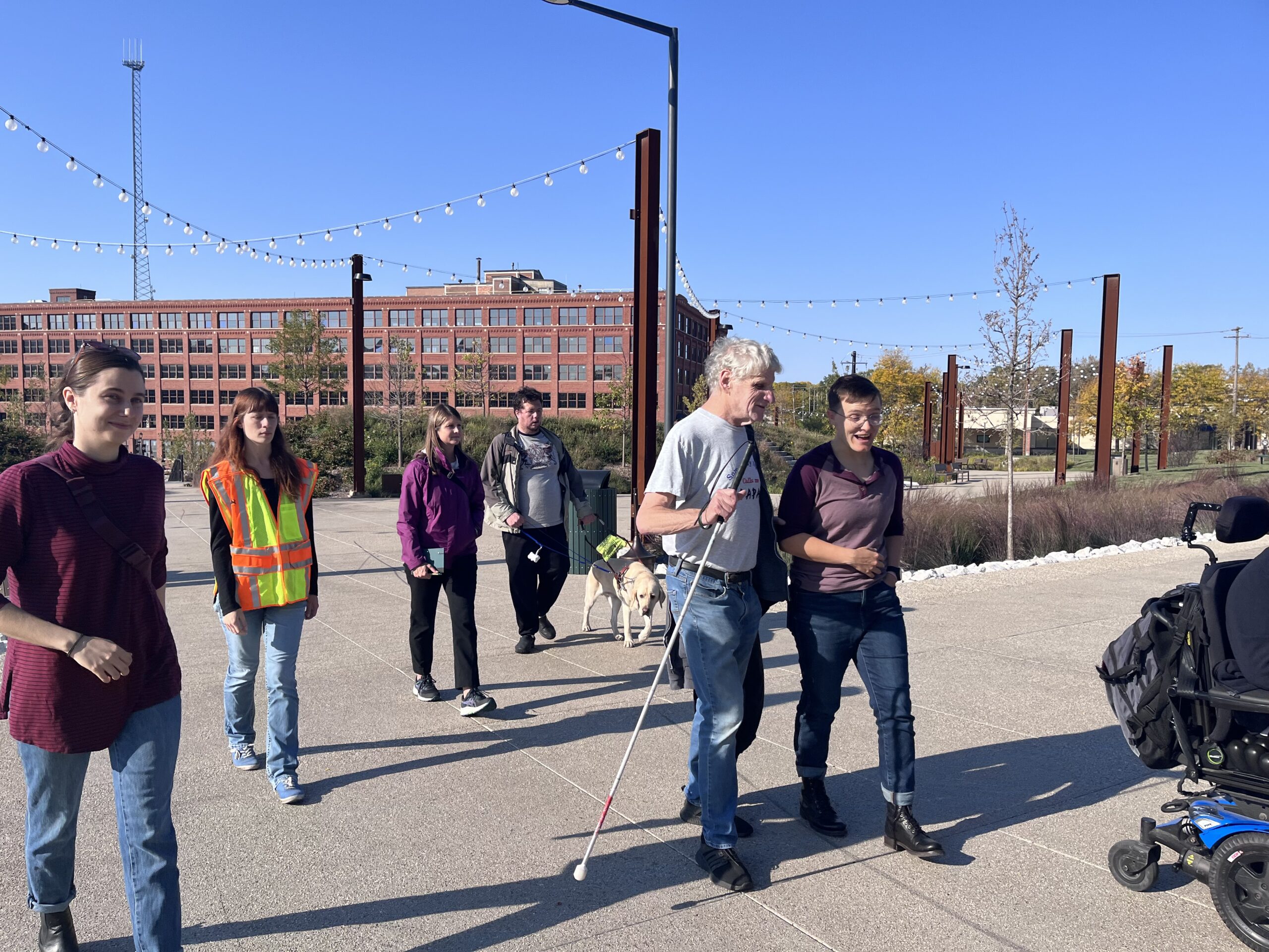 A group of people walk in Davidson Park. In the foreground, a man with a white cane holds the arm of a woman as they talk. In the background are various people. There is a power wheelchair in the foreground although the user is not visible.