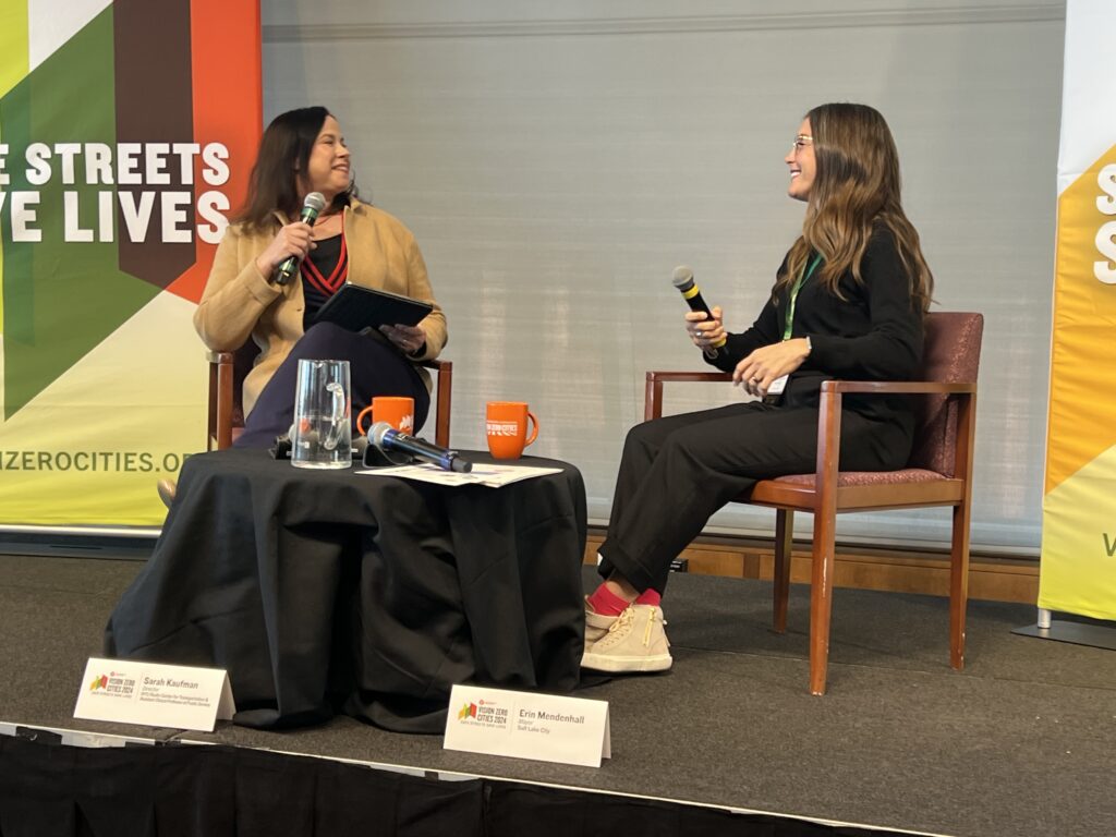 Two women sit on a conference stage, smiling at each other and holding microphones. 